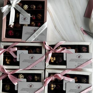Puede incluir: Varias cajas de bombones variados, cada una con una cinta rosa o blanca y una tarjeta. Los bombones est&aacute;n dispuestos en filas e incluyen varios ingredientes. Las cajas tienen el texto "Petal & Cocoa".