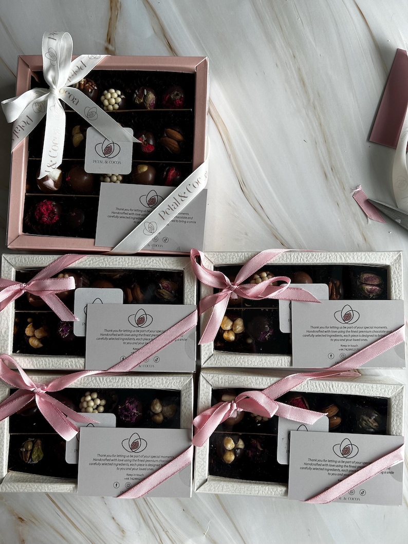 Puede incluir: Varias cajas de bombones variados, cada una con una cinta rosa o blanca y una tarjeta. Los bombones est&aacute;n dispuestos en filas e incluyen varios ingredientes. Las cajas tienen el texto "Petal & Cocoa".