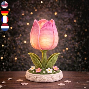 Könnte beinhalten: Eine gehäkelte Tulpenlampe mit rosa Blütenblättern und einem grünen Stiel und Blättern. Die Lampe steht auf einem weißen Sockel, der mit kleinen Blumen verziert ist. Die Lampe wird von innen beleuchtet und erzeugt ein warmes Leuchten.