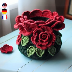 Könnte beinhalten: Ein gehäkelter Blumentopf mit grünem Sockel und roten Rosen. Der Topf ist mit grünen Blättern verziert.