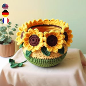 Op de afbeelding: Een gehaakte zonnebloemmand met groene bladeren. De mand is gemaakt van geel en groen garen en heeft drie grote zonnebloemen.