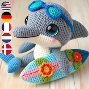Könnte beinhalten: Ein grauer gehäkelter Delfin mit blauer Sonnenbrille, der ein buntes Surfbrett mit Blumen hält. Der Delfin ist ein handgemachtes Spielzeug.