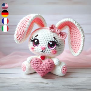 May include: A crocheted pink and white bunny rabbit holding a pink heart. The bunny has large eyes with pink glitter and long eyelashes. The bunny is wearing a pink bow.