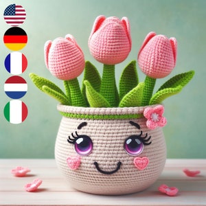 May include: A crocheted flower pot with a smiling face, pink flowers, and green leaves. The pot has large eyes, eyelashes, and pink cheeks.