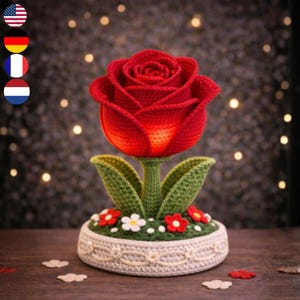 Op de afbeelding: Een handgemaakte gehaakte roos in levendige rode en oranje tinten, met groene bladeren en stengel. De bloem staat in een witte pot versierd met kleine rode en witte bloemen, een charmant decoratief stuk.