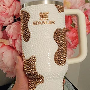 May include: A white Stanley tumbler with a cow print design made of gold-colored rhinestones. The tumbler has a white handle and lid, and a clear straw. The Stanley logo is visible in gold. The background features pink flowers.