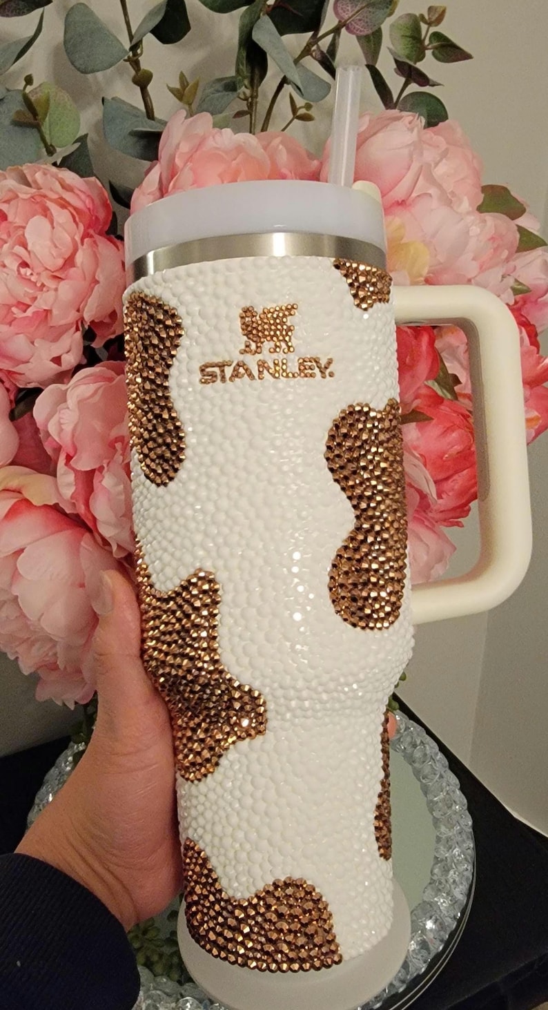 May include: A white Stanley tumbler with a cow print design made of gold-colored rhinestones. The tumbler has a white handle and lid, and a clear straw. The Stanley logo is visible in gold. The background features pink flowers.