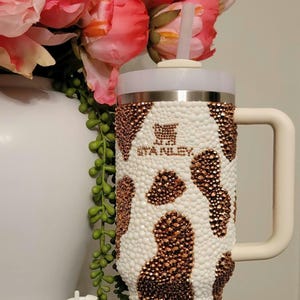 May include: A white and brown bedazzled Stanley tumbler with a matching keychain. The tumbler has a handle and a straw. The design resembles a cowhide pattern. The tumbler and keychain are decorated with small, sparkling embellishments.