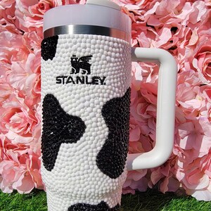 May include: A white and black Stanley tumbler with a cow print design. The tumbler has a white handle and a clear straw. The Stanley logo is visible on the front of the tumbler. The tumbler is set against a backdrop of pink flowers and green grass.