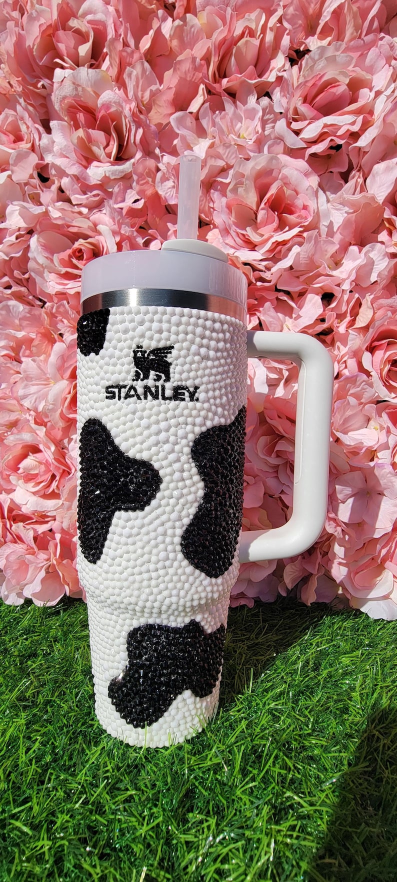 May include: A white and black Stanley tumbler with a cow print design. The tumbler has a white handle and a clear straw. The Stanley logo is visible on the front of the tumbler. The tumbler is set against a backdrop of pink flowers and green grass.