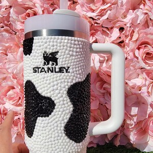 May include: A white and black cow-print Stanley tumbler with a white handle and lid. The tumbler has a straw and the Stanley logo. The background is a pink floral arrangement.