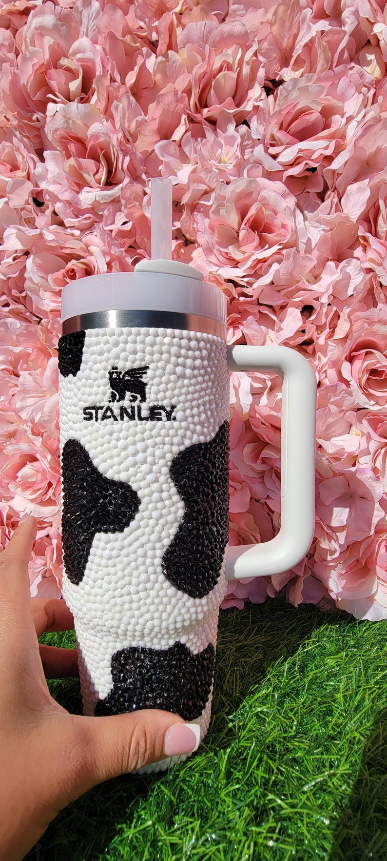 May include: A white and black cow-print Stanley tumbler with a white handle and lid. The tumbler has a straw and the Stanley logo. The background is a pink floral arrangement.
