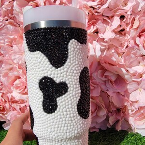 May include: A white and black cow print tumbler with a straw. The tumbler is covered in small, white and black beads. The background is pink flowers and green grass.