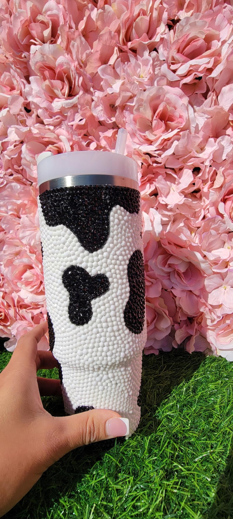 May include: A white and black cow print tumbler with a straw. The tumbler is covered in small, white and black beads. The background is pink flowers and green grass.