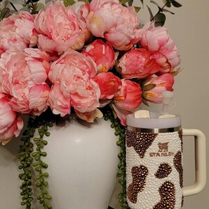 May include: A white vase filled with pink peonies and greenery. A bedazzled Stanley tumbler with a cow print design and a matching keychain tumbler are next to the vase. The tumblers have a white and brown pattern.