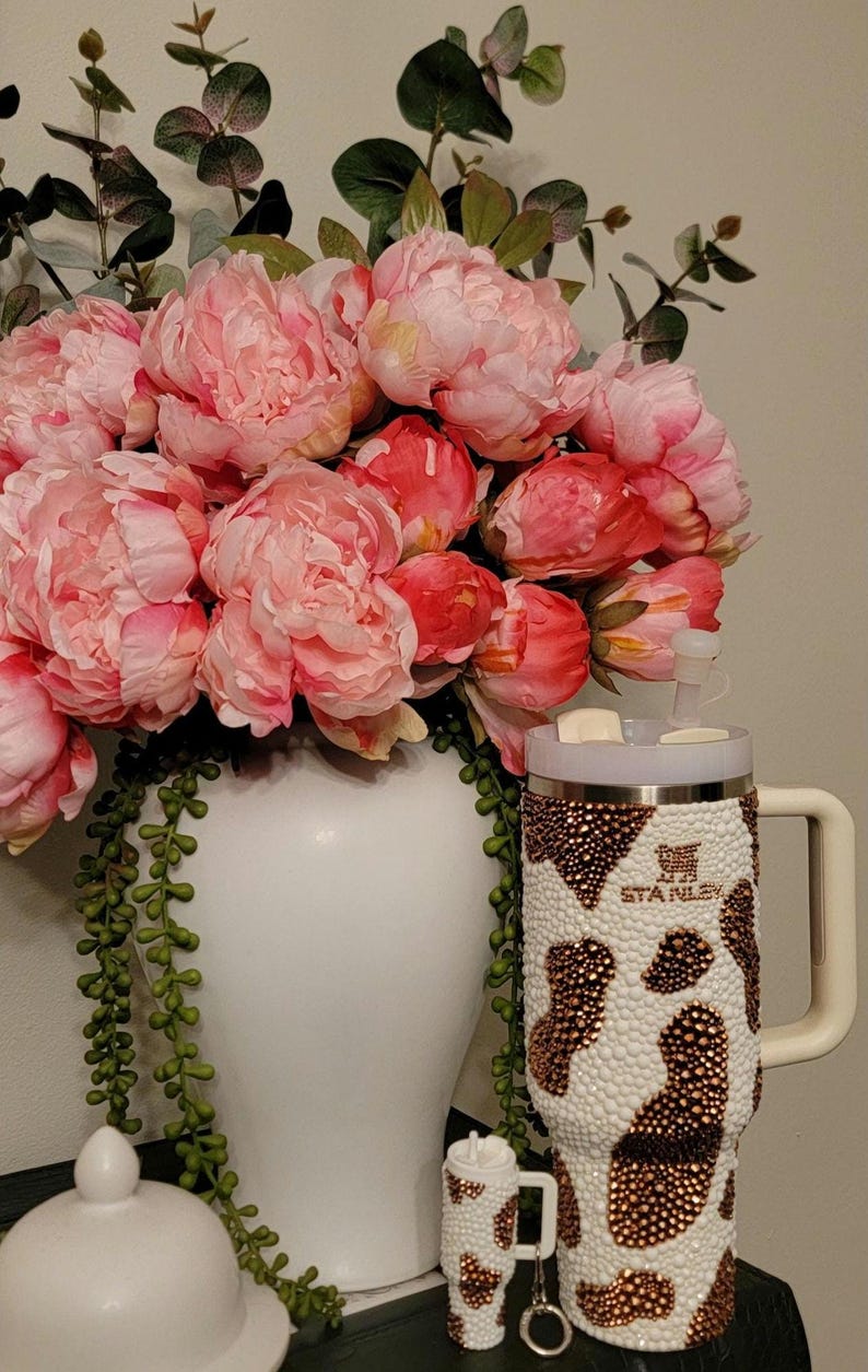 May include: A white vase filled with pink peonies and greenery. A bedazzled Stanley tumbler with a cow print design and a matching keychain tumbler are next to the vase. The tumblers have a white and brown pattern.