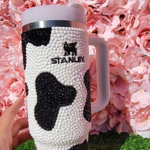 May include: A Stanley tumbler with a white handle and straw, decorated with a cow print pattern of black and white rhinestones. The tumbler has the Stanley logo and is set against a backdrop of pink flowers.