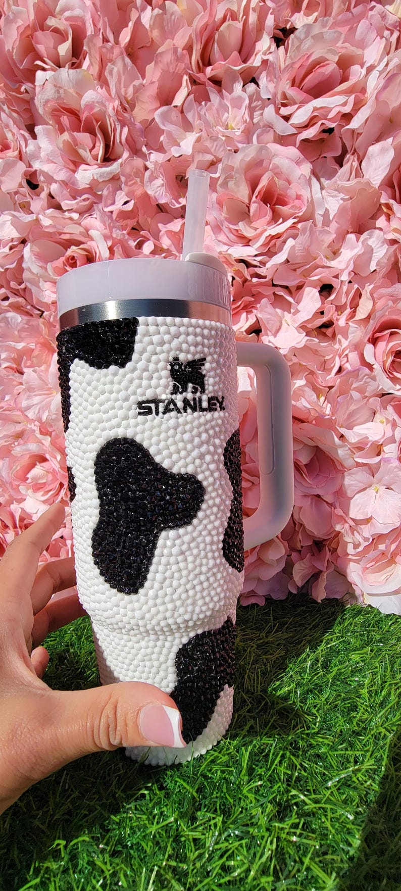 May include: A Stanley tumbler with a white handle and straw, decorated with a cow print pattern of black and white rhinestones. The tumbler has the Stanley logo and is set against a backdrop of pink flowers.