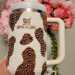 May include: A white and brown bedazzled Stanley tumbler with a handle and straw. The tumbler has a cow print design with the brand name "STANLEY" in the center. The tumbler is held in a person's hand.