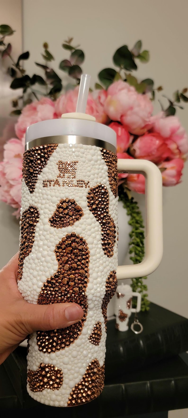 May include: A white and brown bedazzled Stanley tumbler with a handle and straw. The tumbler has a cow print design with the brand name "STANLEY" in the center. The tumbler is held in a person's hand.