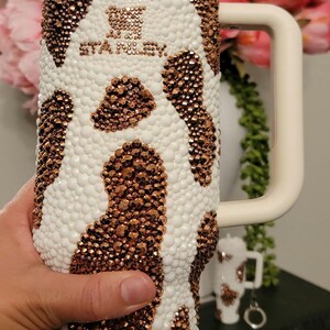 May include: White and copper-colored bedazzled tumbler with a handle and straw. The tumbler has a cow-like pattern with the text "STANLEY" on the front. The tumbler is decorated with small, shiny embellishments.