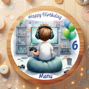 Könnte beinhalten: Ein runder Kuchen, der mit einer Cartoon-Illustration eines Jungen mit Kopfhörern und Videospielen dekoriert ist. Der Kuchen ist mit den Worten "Happy Birthday" und "Manu" sowie der Zahl "6" verziert.