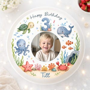 Può includere: Un piatto in ceramica bianco con un colorato design a tema sottomarino. Il piatto presenta una foto di un bambino sorridente al centro, circondata da illustrazioni di creature marine, il numero 3 e le parole "Happy Birthday" e il nome "Till".