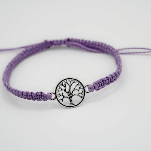 Pode incluir: Uma pulseira de macramê lavanda com um berloque de árvore da vida prateado. A pulseira é ajustável e apresenta um design trançado. O berloque de árvore é uma silhueta detalhada dentro de uma moldura circular.