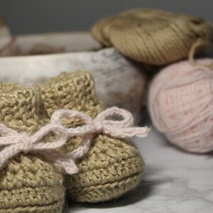 Könnte beinhalten: Ein Paar gehäkelte Babyschuhe in beige mit rosa Schleifen. Die Schuhe liegen auf einer weißen Marmorfläche.