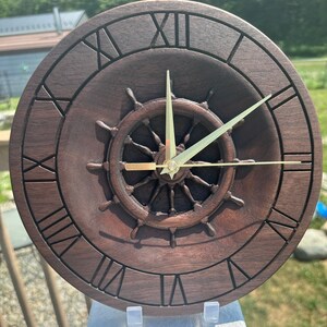 May include: A dark brown wooden clock with Roman numeral markings and a carved ship's wheel design. The clock hands are gold.