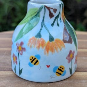 May include: A hand-painted ceramic vase with a light blue base. The vase features orange and yellow flowers, green leaves, and cartoon bees. The design includes small pink flowers and a red heart. The vase is on a wooden surface.