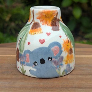 May include: A small, white ceramic vase with a hand-painted design. The vase features a blue koala bear, orange flowers, purple flowers, green leaves, and red hearts. The vase is sitting on a wooden surface.