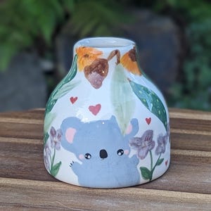 May include: A white ceramic vase featuring a koala design. The vase is decorated with green leaves, orange flowers, and red hearts. The koala is light blue with a white belly and black nose.