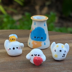 May include: A collection of quirky ceramic figurines. The set features a small vase with a blue cloud design and several ghost-like figures, one holding a red heart. The figurines are hand-painted with bright colours and playful details.