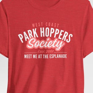 Puede incluir: Una camiseta roja de manga corta con texto en blanco y rosa claro que dice West Coast Park Hoppers Society Estd. 2001 Meet Me At The Esplanade. La camiseta tiene un cuello redondo y una textura jaspeada en toda la tela.