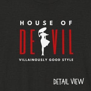 Shown is the detailed view of the design. Top line of reads HOUSE OF, followed by the word DE VIL, with the silhouette of a woman in between the E and the V. Below are the words Villainously Good Style. Shown is the Solid White Blend shirt color option.