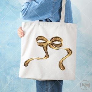 May include: A white tote bag featuring a gold glitter bow design. The bow is large and has a three-dimensional appearance.
