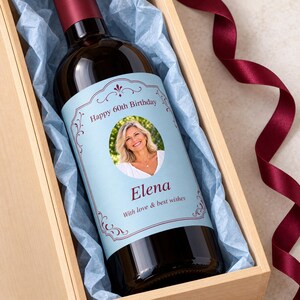Puede incluir: Una botella de vino de vidrio oscuro con una etiqueta azul claro personalizada que muestra una foto circular de una persona sonriente, el texto Happy 60th Birthday, el nombre Elena y la frase With love & best wishes, en una caja de madera.