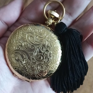 May include: A gold-colored pocket watch with an ornate floral design on the cover. The watch has a gold ring and a black tassel attached. The watch is round and appears to be vintage.