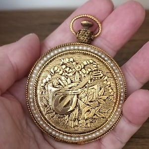 May include: A gold-coloured pocket watch pendant with an embossed design of musical instruments and flowers. The watch is edged with small, white pearls. It has a loop at the top for suspension. The pendant is circular in shape.