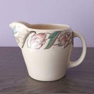 Peut inclure: Crémier en céramique crème avec un motif floral. Le crémier a un bec verseur et une anse. Le motif floral présente des fleurs et des feuilles roses, vertes et grises. Le crémier est posé sur une surface en bois.