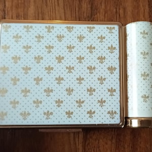 May include: A vintage compact and lipstick set. The set has a light blue background with a gold fleur-de-lis pattern. The compact is rectangular with a gold frame, and the lipstick has a matching design.