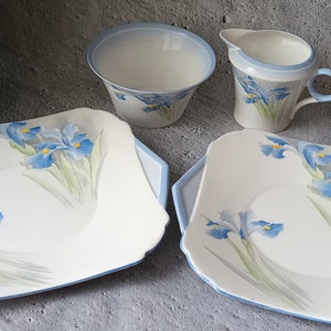 May include: Two square white ceramic cake plates with blue trim and a blue and green floral design of irises. A white ceramic creamer and sugar bowl with blue trim are also included.