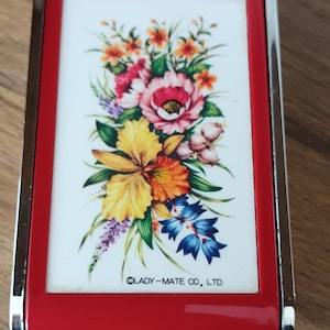 May include: A rectangular, red compact mirror with a silver trim. The mirror features a floral design with pink, yellow, orange, and blue flowers on a white background. The text "LADY-MATE CO., LTD" is printed at the bottom.