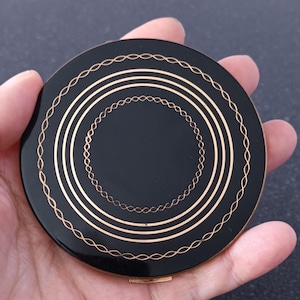 May include: A black compact with gold-colored decorative lines and a gold clasp. The compact has a circular design with concentric circles and a delicate chain-like pattern around the edges. The compact is held in a hand.