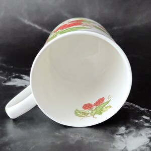 K&ouml;nnte beinhalten: Wei&szlig;e Keramiktasse mit rotem und gr&uuml;nem Blumendesign. Die Tasse hat einen Henkel und ist leer.