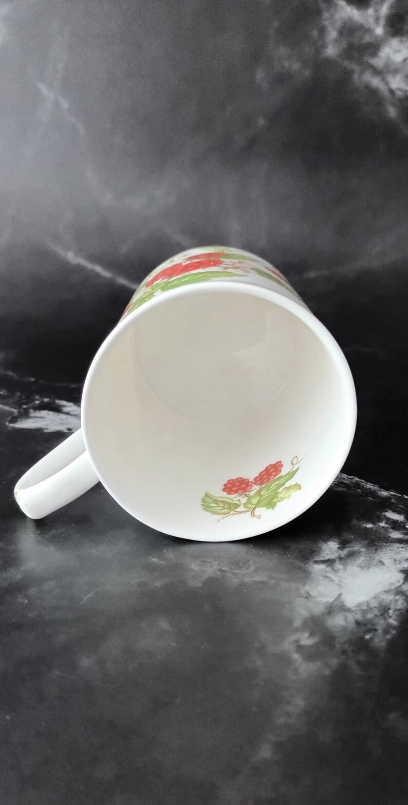 K&ouml;nnte beinhalten: Wei&szlig;e Keramiktasse mit rotem und gr&uuml;nem Blumendesign. Die Tasse hat einen Henkel und ist leer.