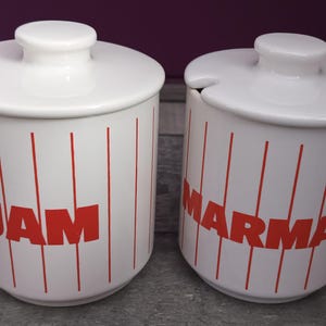 Könnte beinhalten: Zwei weiße Keramikgläser mit roten Deckeln und roten Beschriftungen. Das Glas links trägt die Aufschrift "JAM" und das Glas rechts die Aufschrift "MARMALADE".