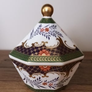 Puede incluir: Un tarro de cerámica blanco con tapa y un adorno dorado. El tarro presenta un diseño geométrico con patrones florales en azul marino, dorado, verde y naranja. El tarro está colocado sobre una superficie de madera.