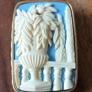 May include: A rectangular brooch featuring a light blue background with a raised cream-colored relief carving. The design depicts a decorative urn filled with leafy foliage, positioned behind a balustrade railing with two tall, slender trees nearby.